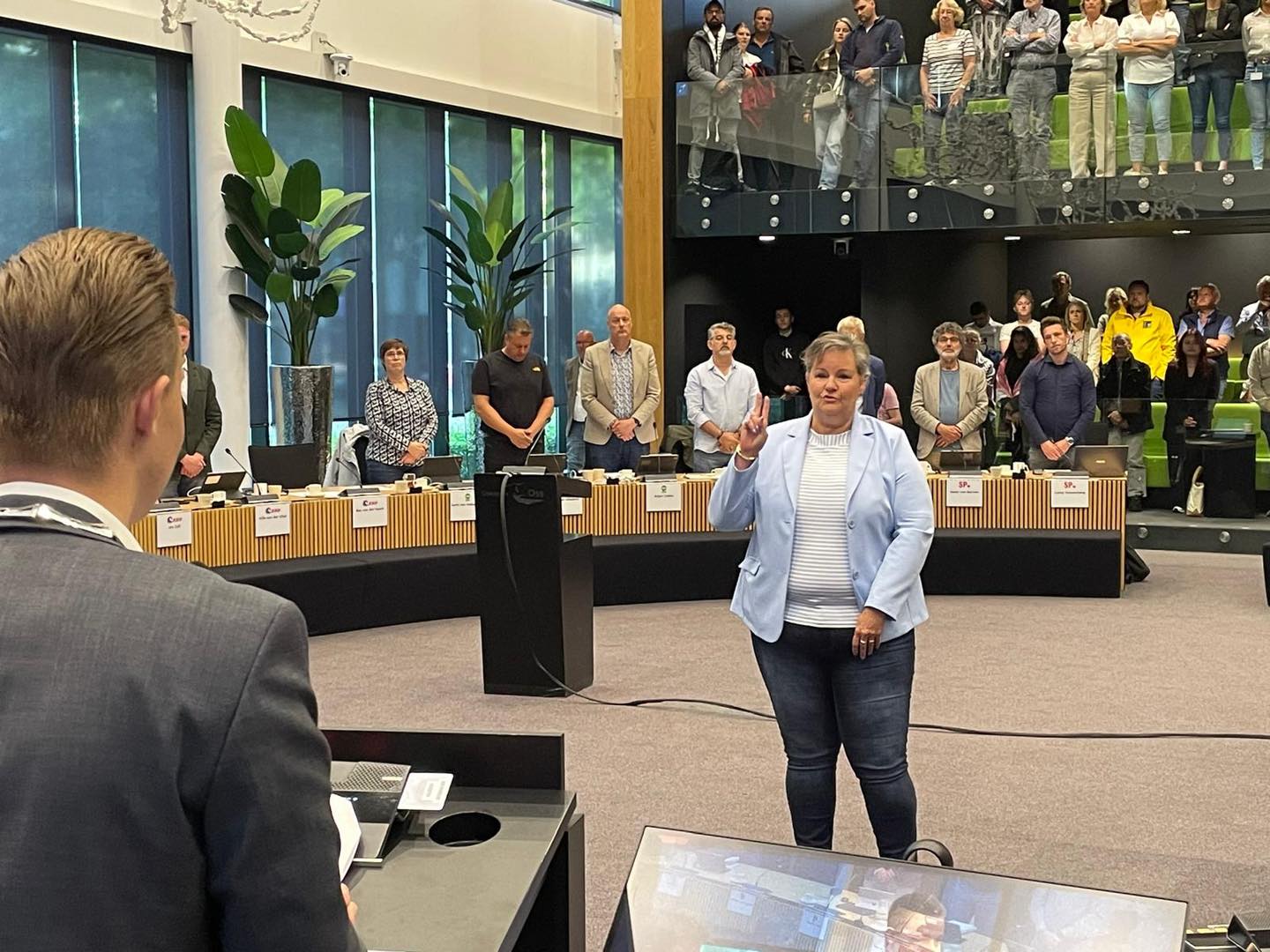 Vrouw maakt soort van eed in vergaderzaal vol staande toeschouwers.