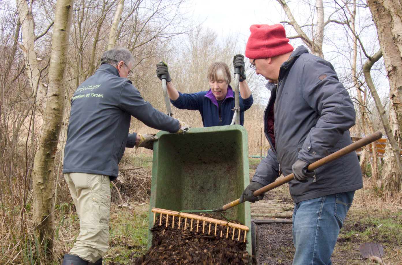 Drie mensen werken samen in een bos, met een kruiwagen en harken om tuinafval te verdelen.