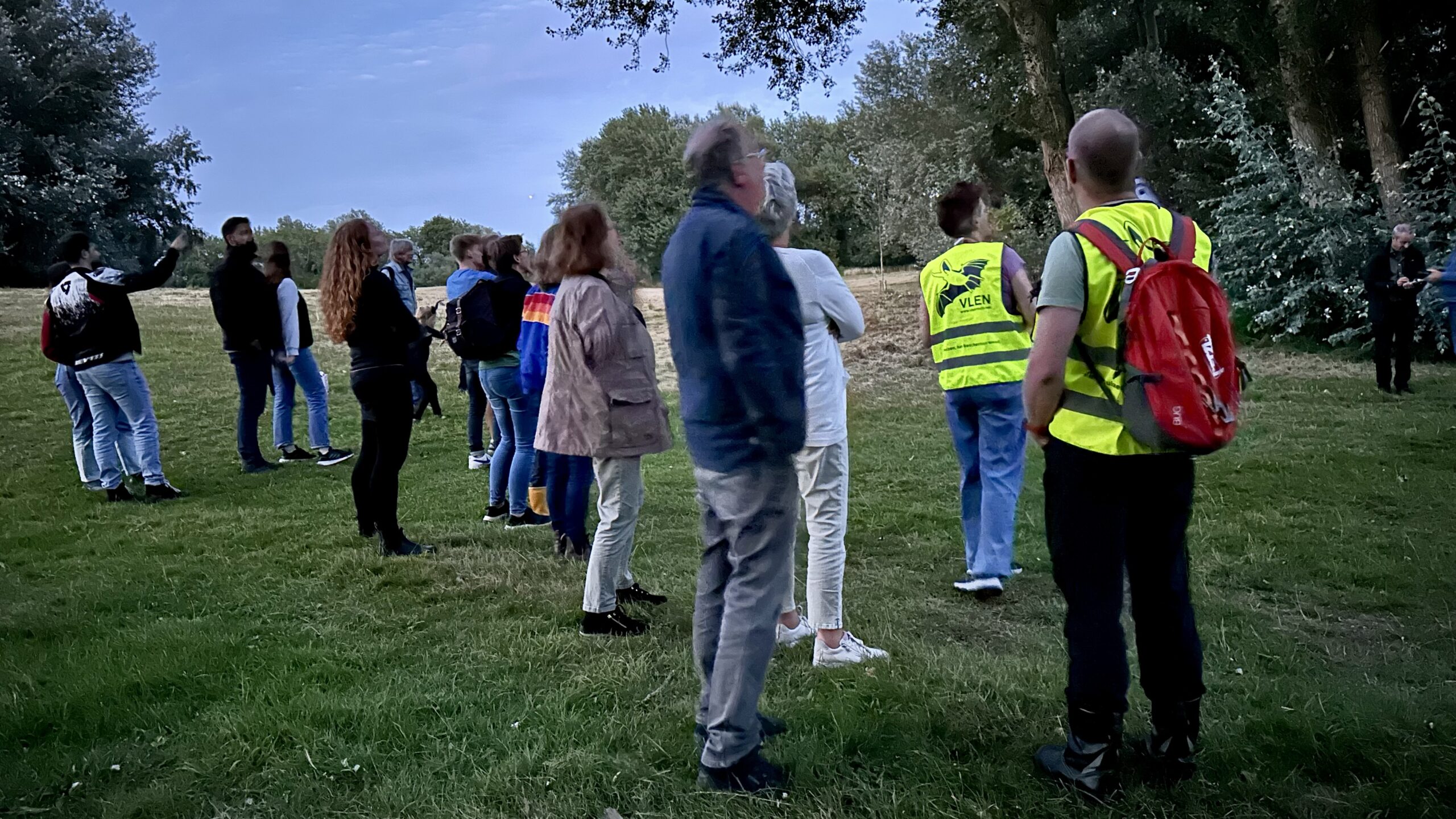 Groep mensen in het gras, sommigen dragen gele hesjes, omgeven door bomen in schemerlicht.