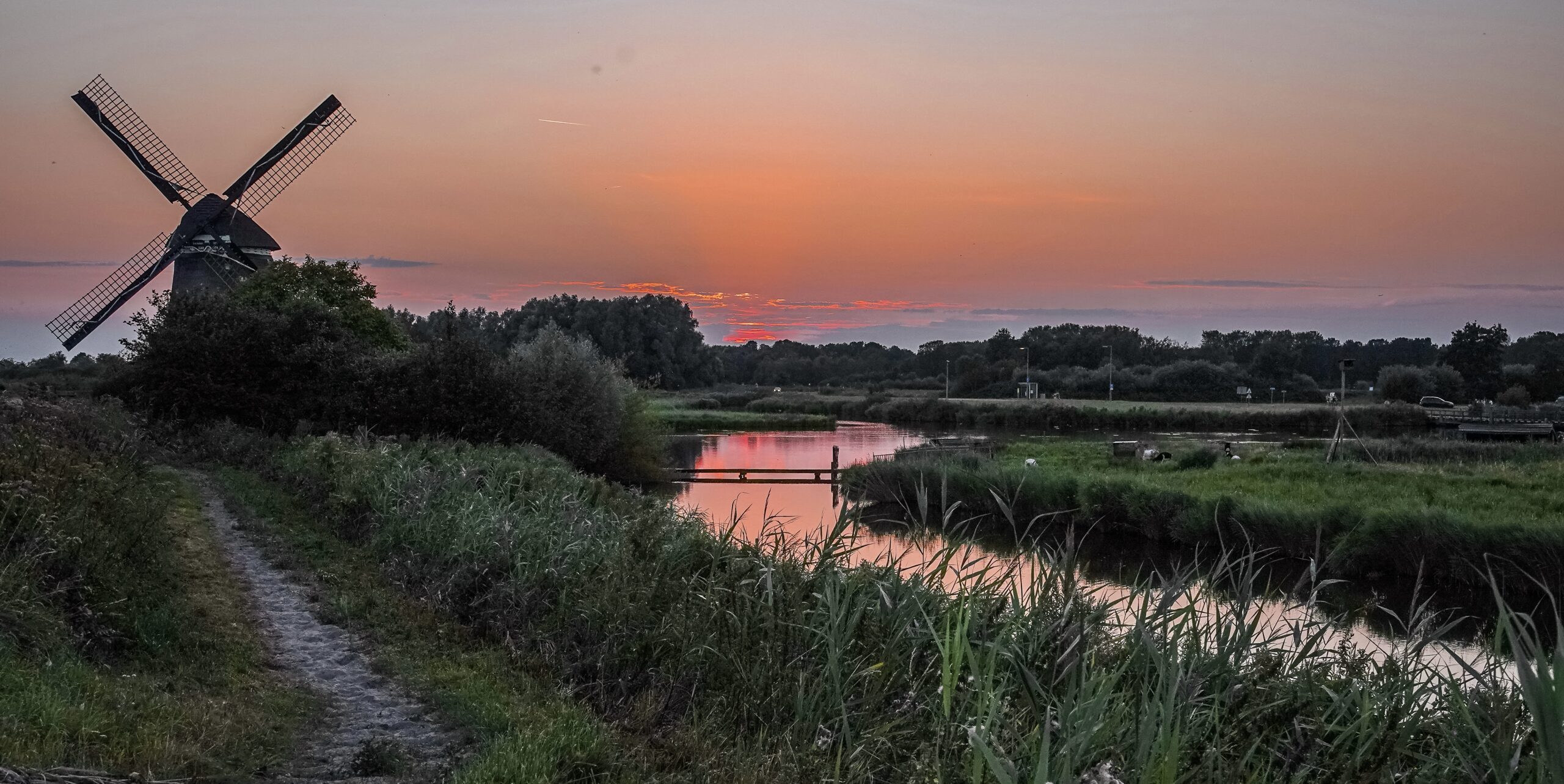 Windmolen bij zonsondergang naast rivier, omgeven door groen landschap.