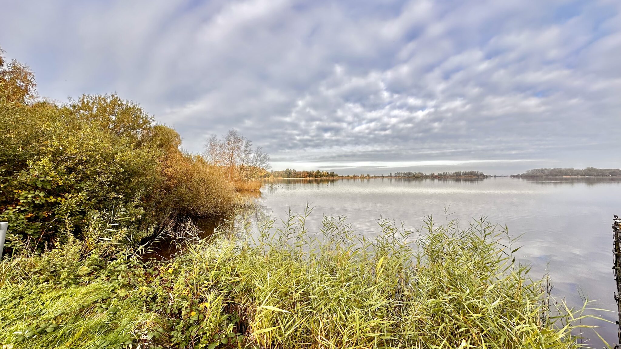 Rustig meer met groene en herfstkleurige begroeiing onder een bewolkte hemel.