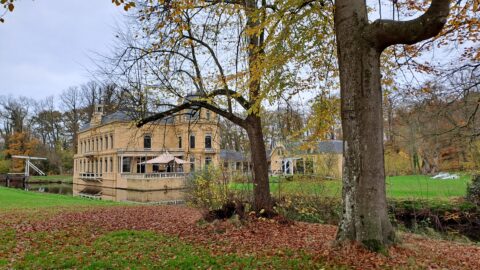 Herfstlandschap met een groot, historisch gebouw bij een gracht, omgeven door bomen en gevallen bladeren.