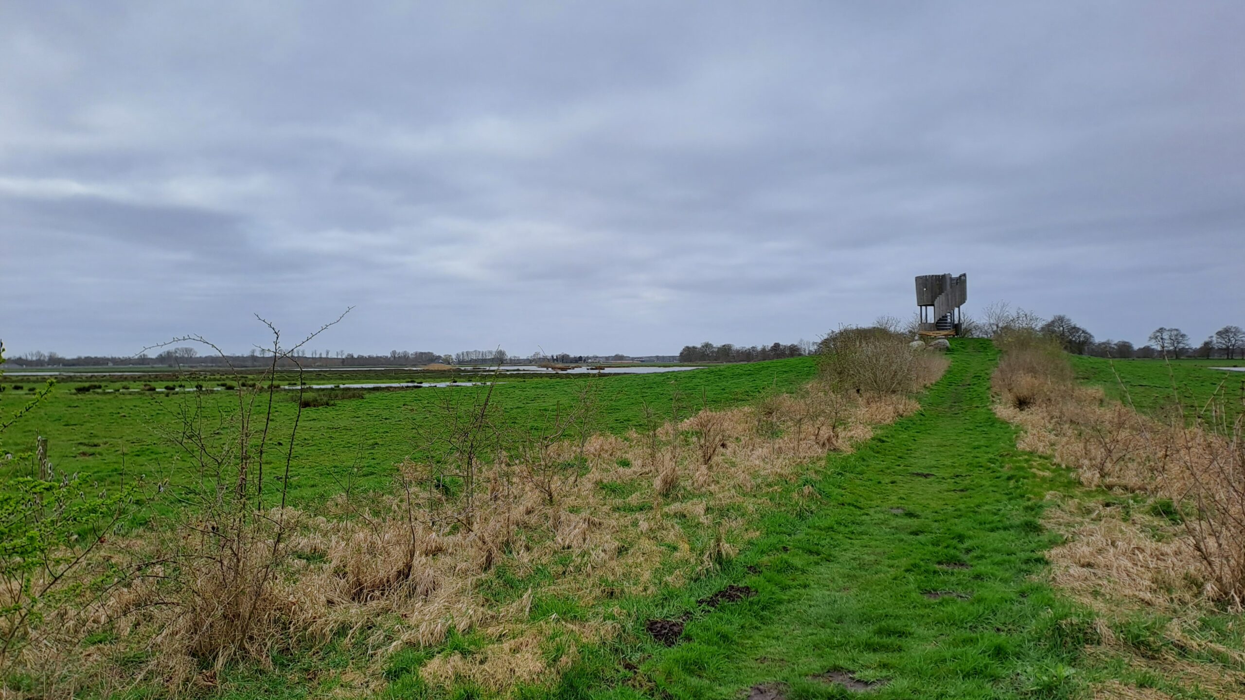Groen veld met pad naar een eenzame houten uitkijktoren onder een bewolkte grijze hemel.