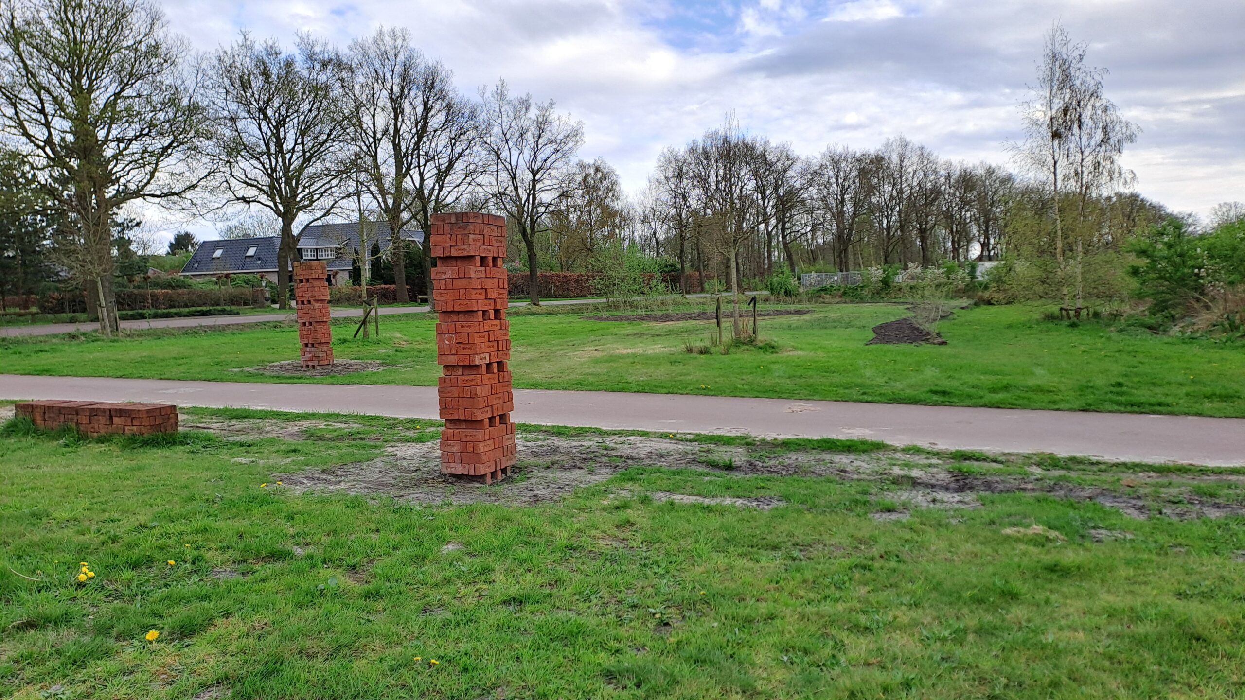 Baksteenzuilen langs een pad in een groen park met kale bomen en een huis op de achtergrond.