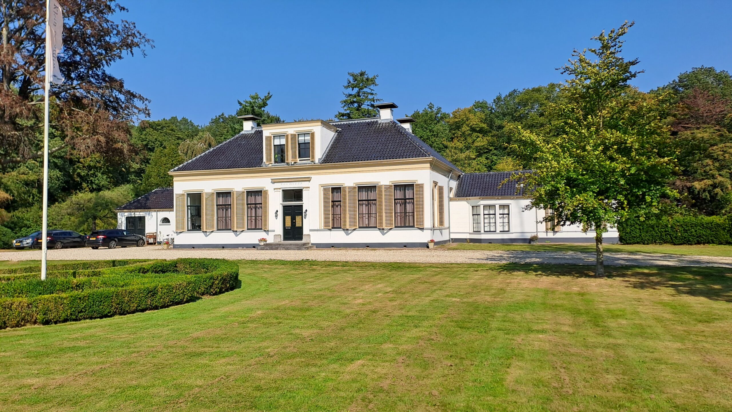 Een statig, wit landhuis met zwarte dakpannen, omgeven door een grasveld en bomen.