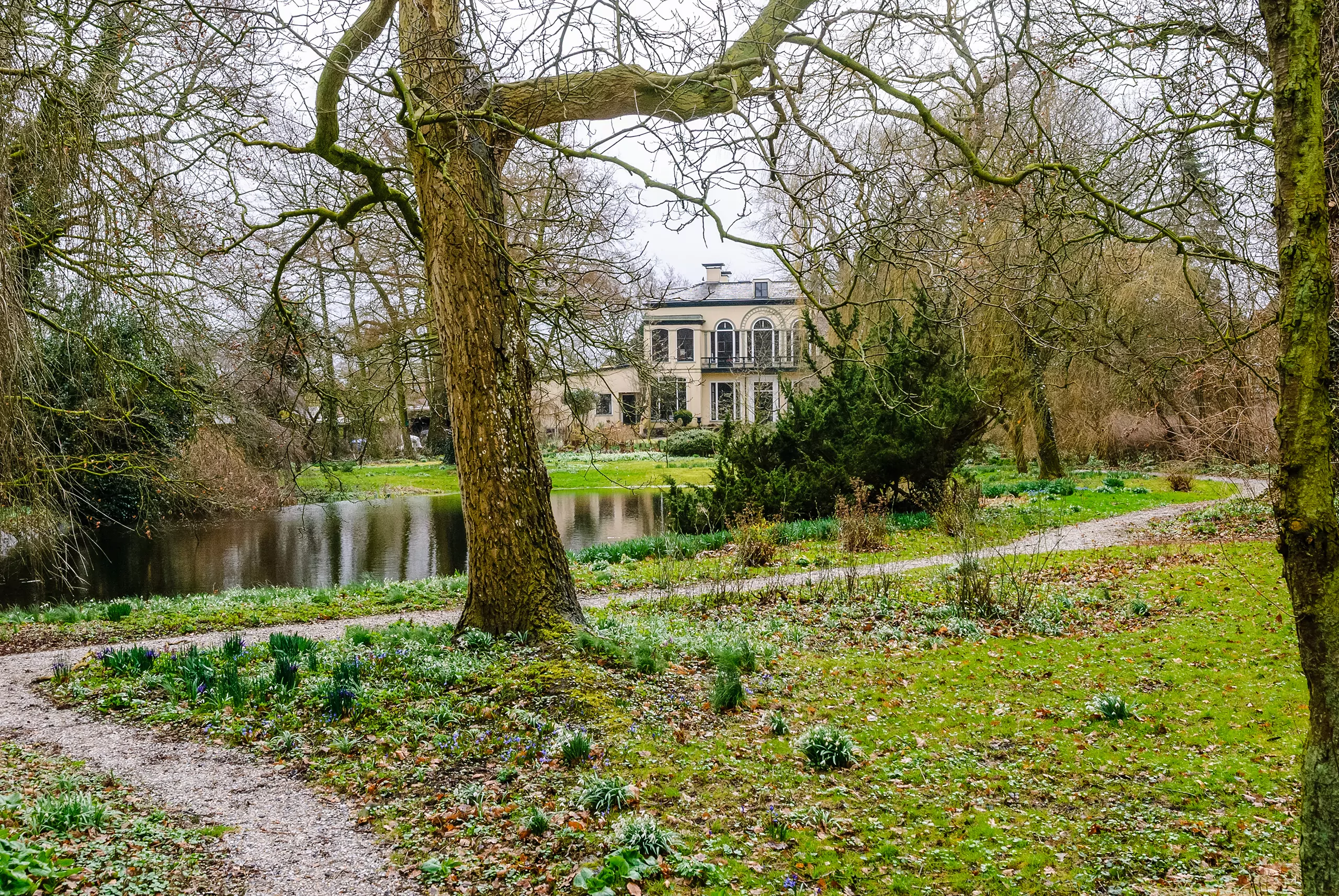 Landhuis omgeven door bomen, paden en een vijver in een groene tuin met bloeiende bloemen.