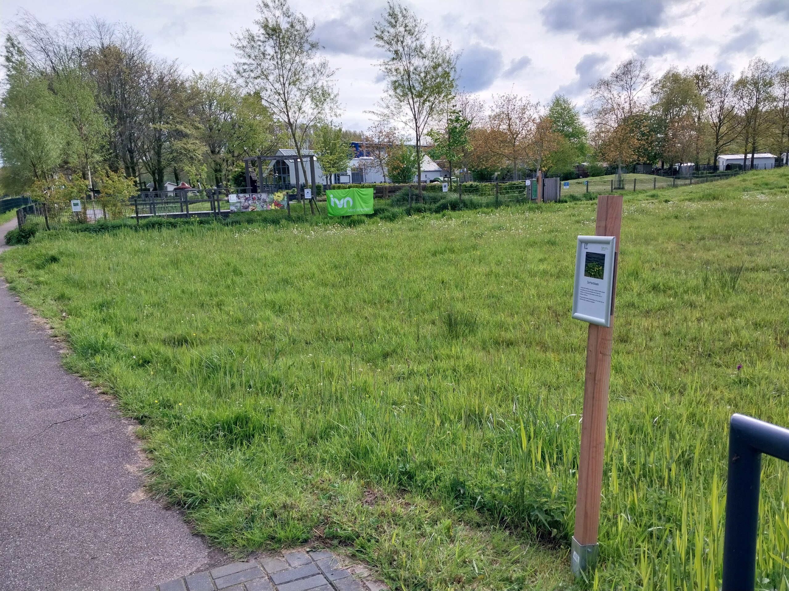 Groen park met een informatiebord en hek. Achteraan staan bomen en een groen IVN-spandoek.