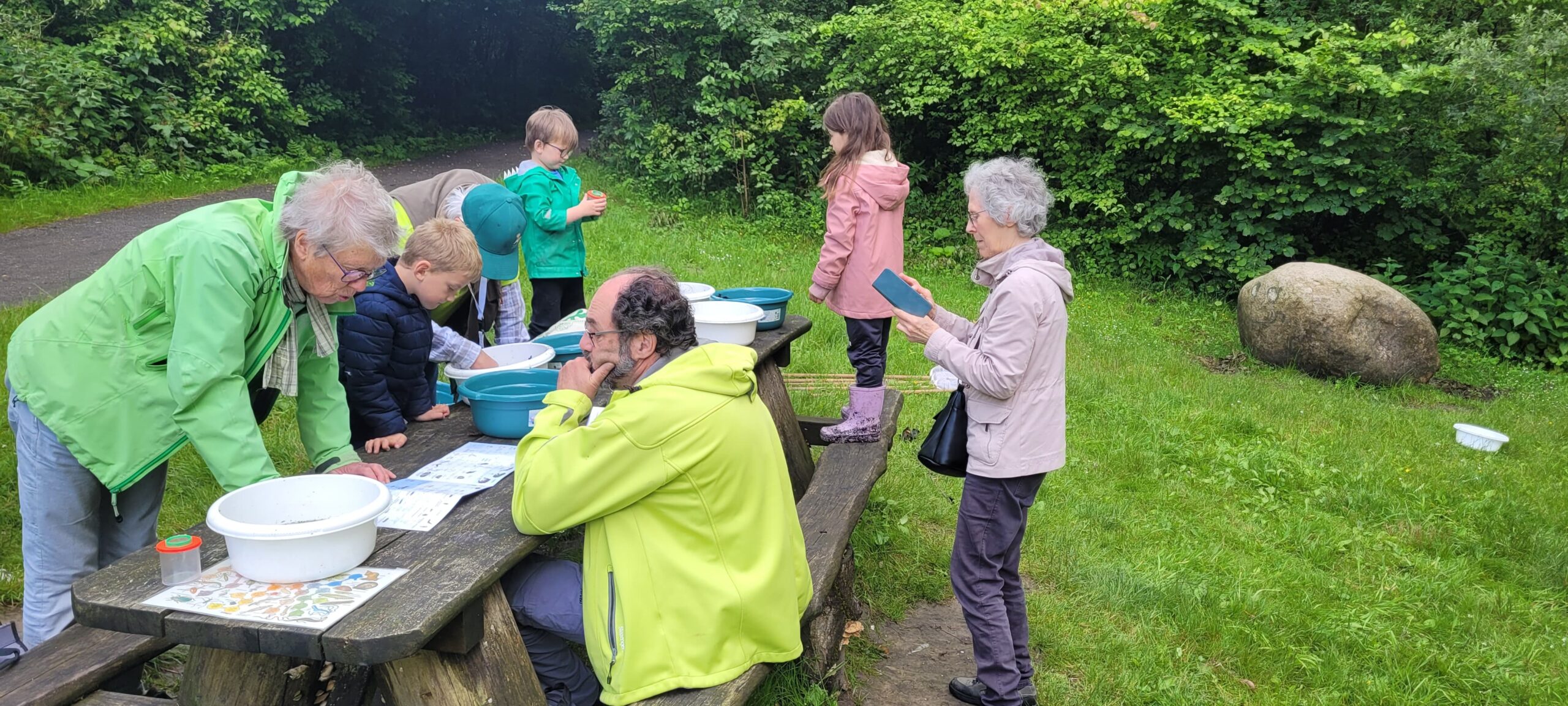 Mensen onderzoeken items aan een picknicktafel in een groene, bosrijke omgeving.