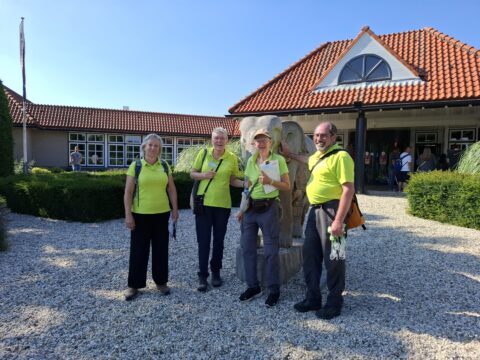 Vier mensen in gele shirts poseren voor een gebouw met rode dakpannen.