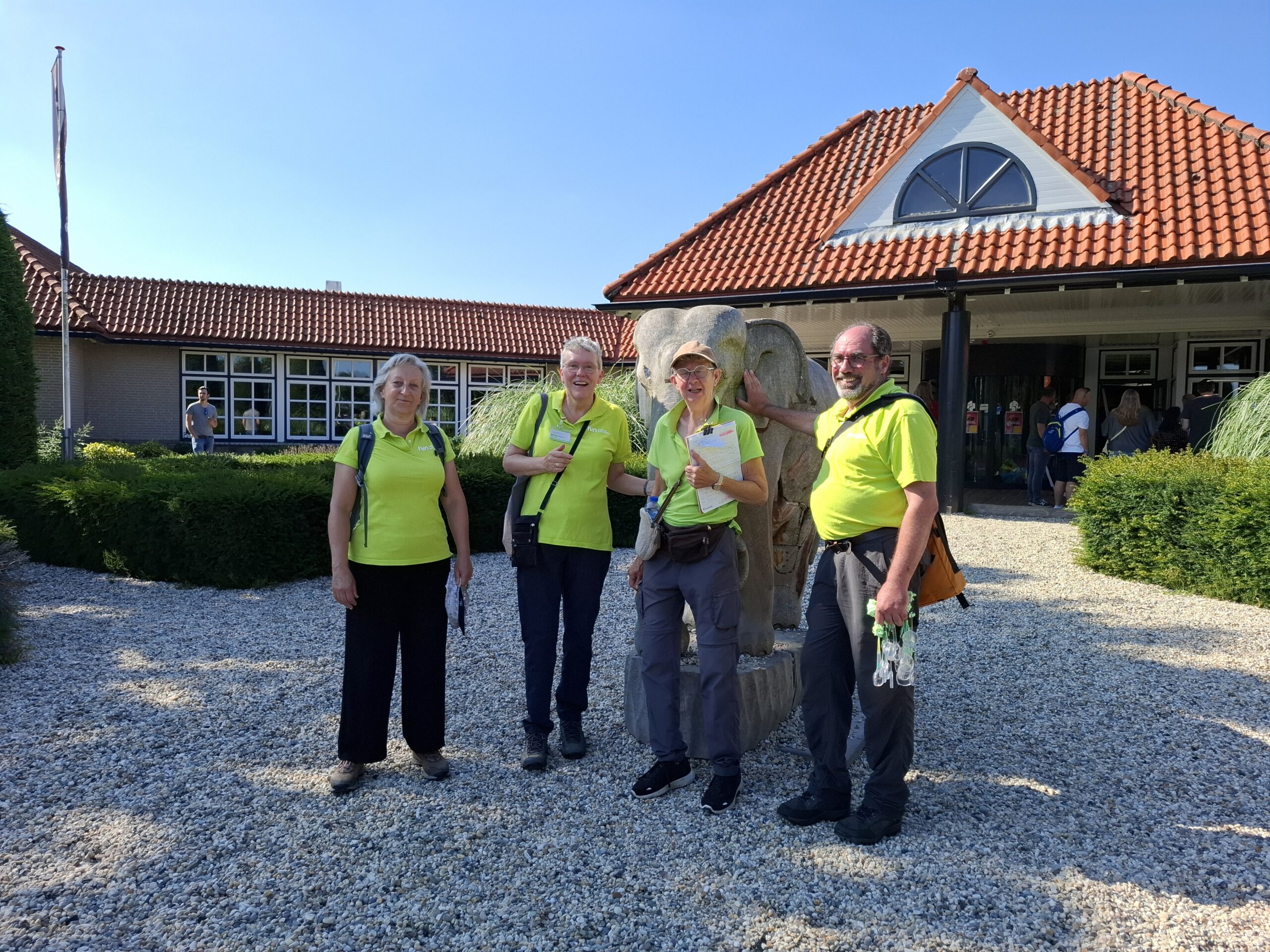 Vier mensen in gele shirts poseren voor een gebouw met rode dakpannen.