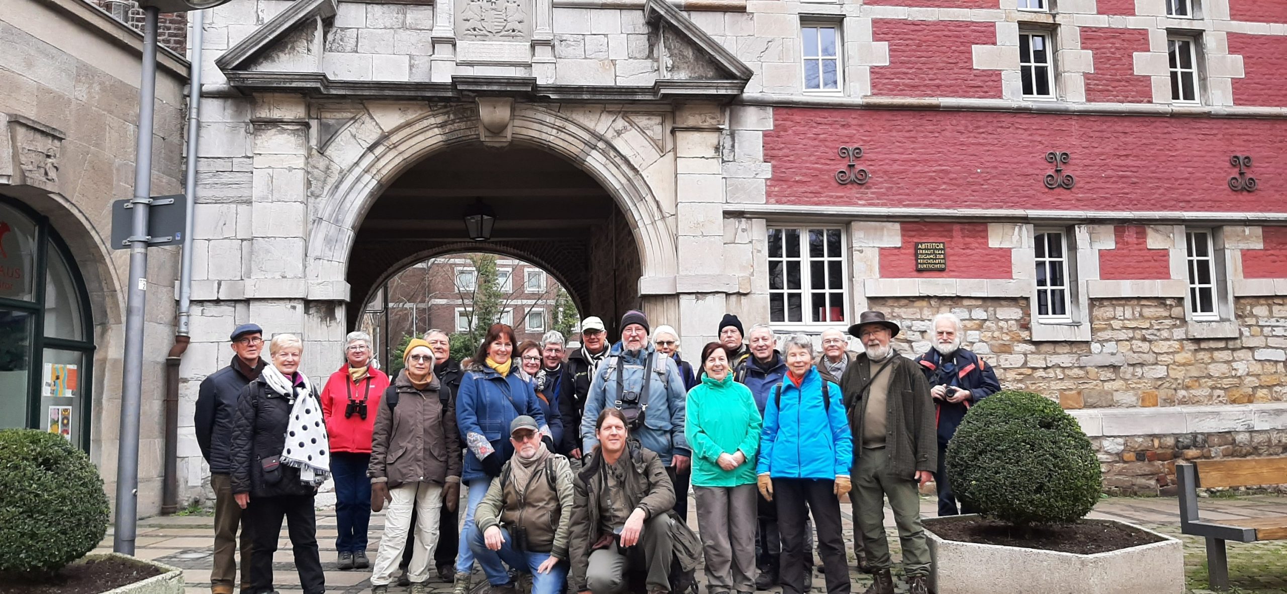 Groep mensen poseert voor een oud stenen gebouw met een boog en rode bakstenen muur.