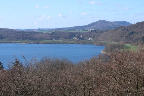 Uitzicht op een meer omgeven door kale bomen, met heuvels en een dorp in de verte onder een blauwe hemel.