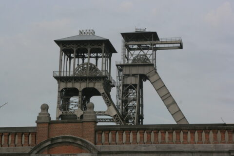 Historische mijnschacht met stalen koeltorens en bakstenen muur op de voorgrond.