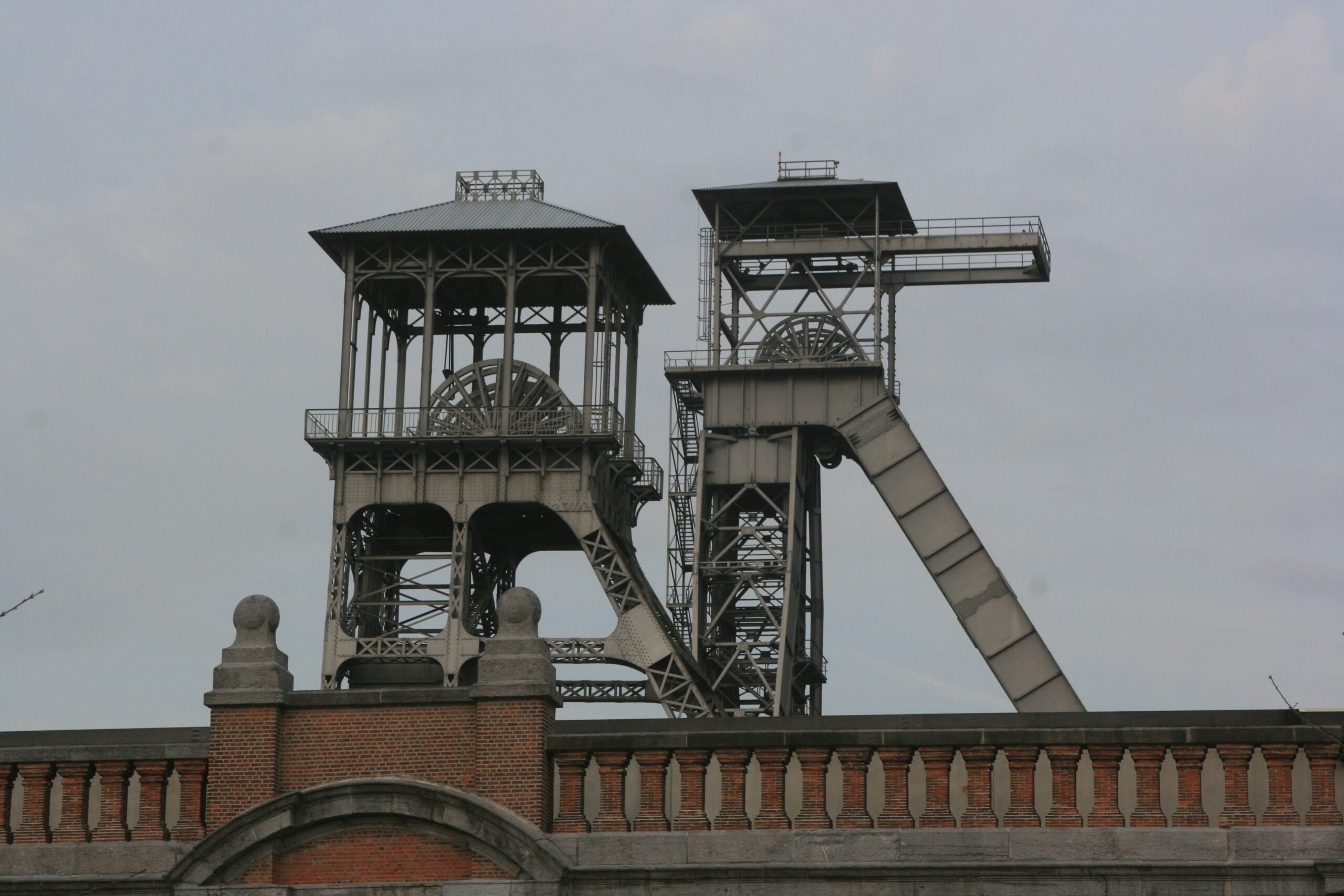 Historische mijnschacht met stalen koeltorens en bakstenen muur op de voorgrond.