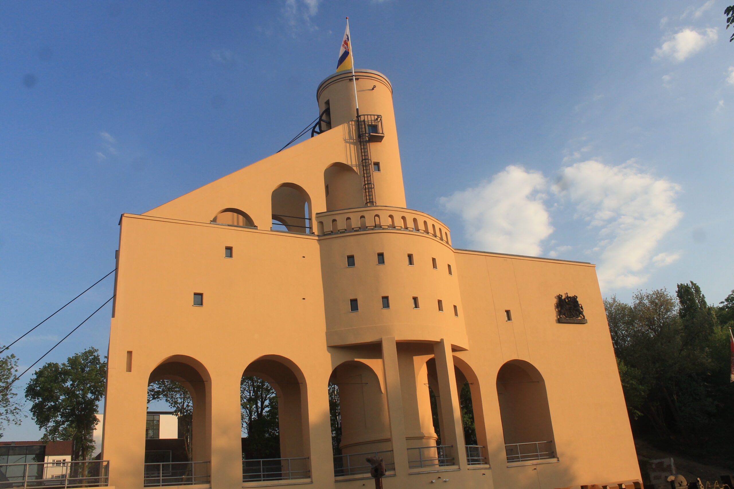 Gele, moderne toren met vlag, omlijst door bogen. Helderblauwe lucht op de achtergrond.