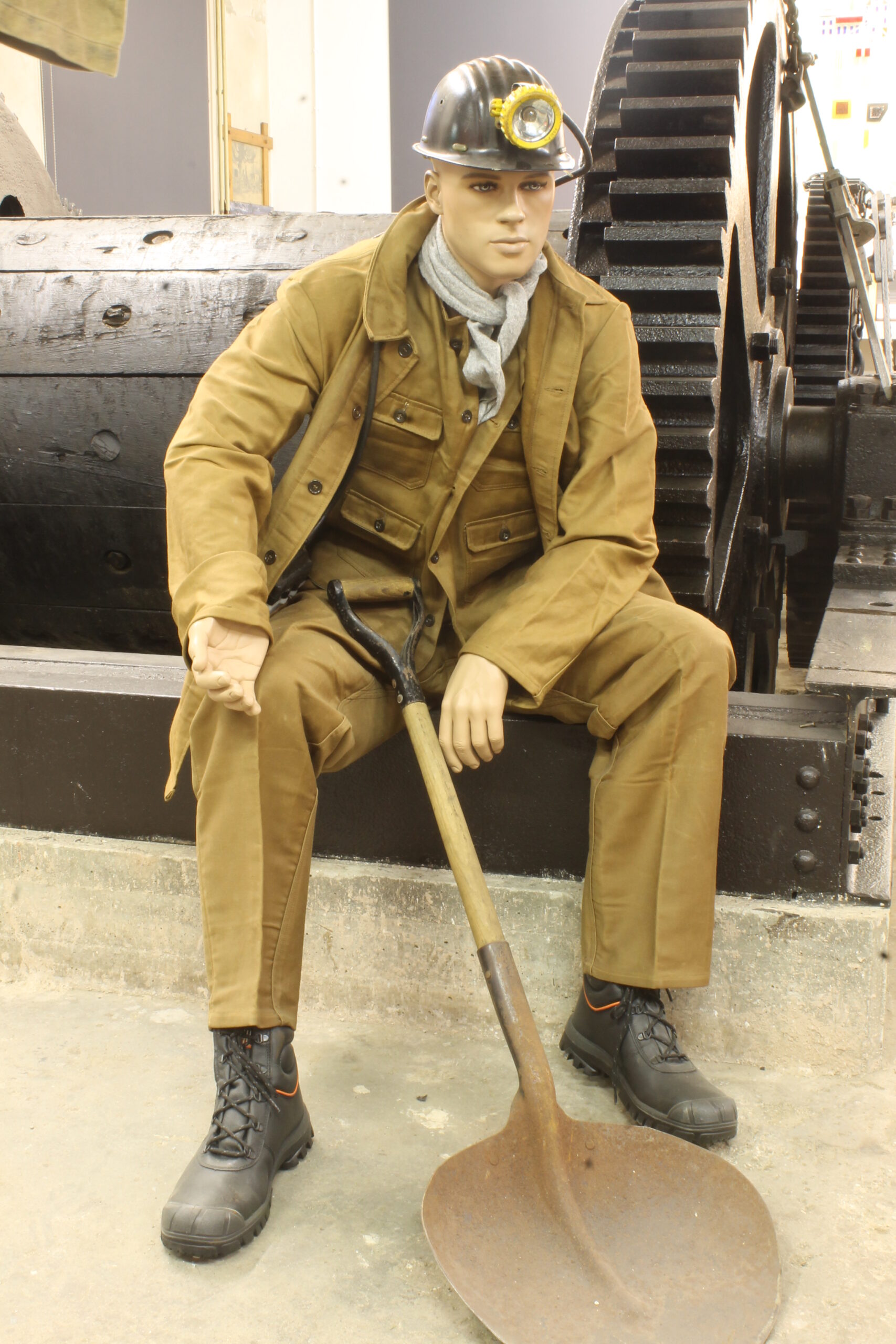 Pop met mijnwerkerskleding, helm en schop, zittend voor grote metalen tandwielen.