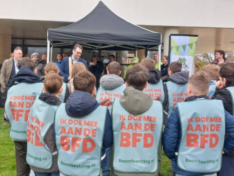 Kinderen in blauwe hesjes verzamelen zich bij een man onder een tent voor Boomfeestdag-presentatie.