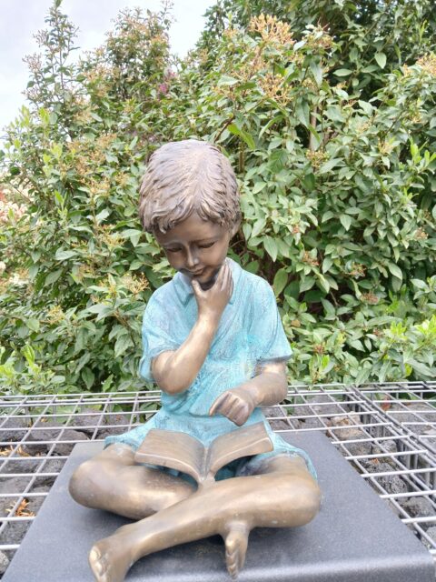 Bronzen standbeeld van een zittend kind dat in een boek leest, omringd door groene struiken.