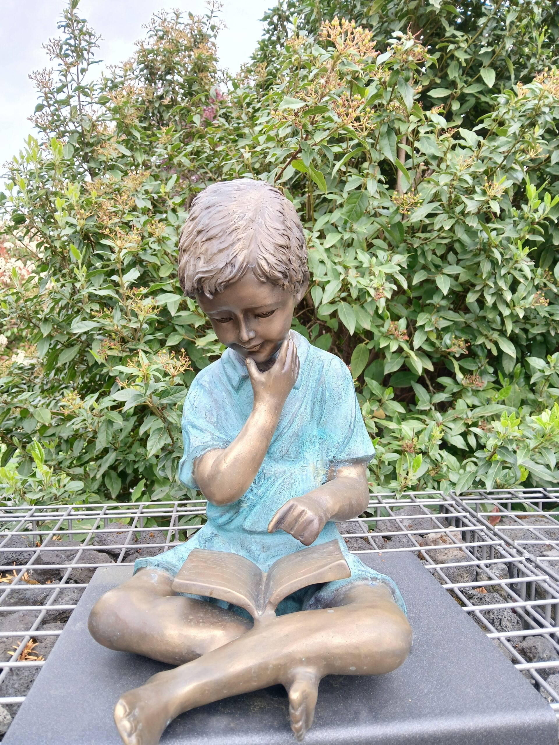 Bronzen standbeeld van een zittend kind dat in een boek leest, omringd door groene struiken.
