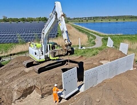Graafmachine plaatst betonnen wandplaten bij zonnepark en meer; persoon in oranje overall kijkt toe.