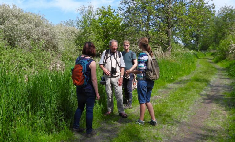 Vier mensen praten op een groen, bosrijk pad met rugzakken en camera's.