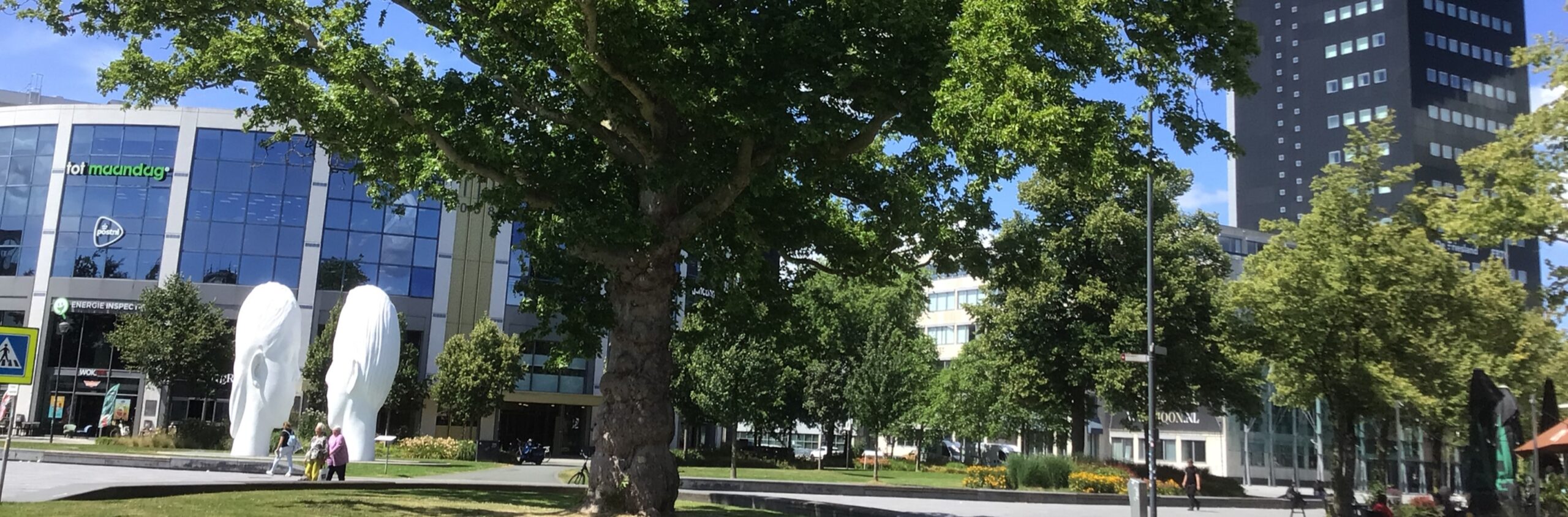 Een park met standbeelden, mensen, bomen, kantoorgebouw en een hoog gebouw op de achtergrond.