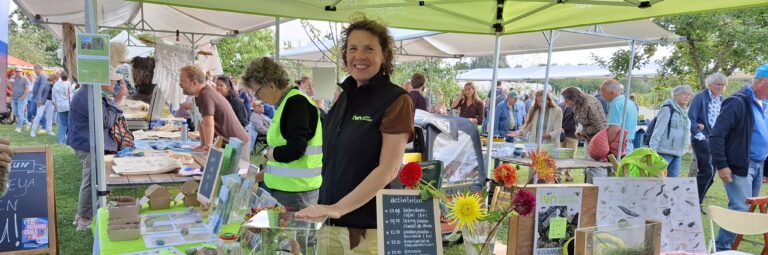 Vrouw bij informatiestand op druk bezochte markt, omringd door bezoekers en bloemen.