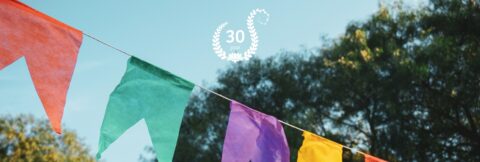 Vlaggetjeslijn in oranje, groen en paars met "30 jaar" in cirkel van bladeren tegen blauwe lucht.