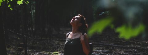Vrouw kijkt omhoog in een schaduwrijk bos, omgeven door groene bladeren.