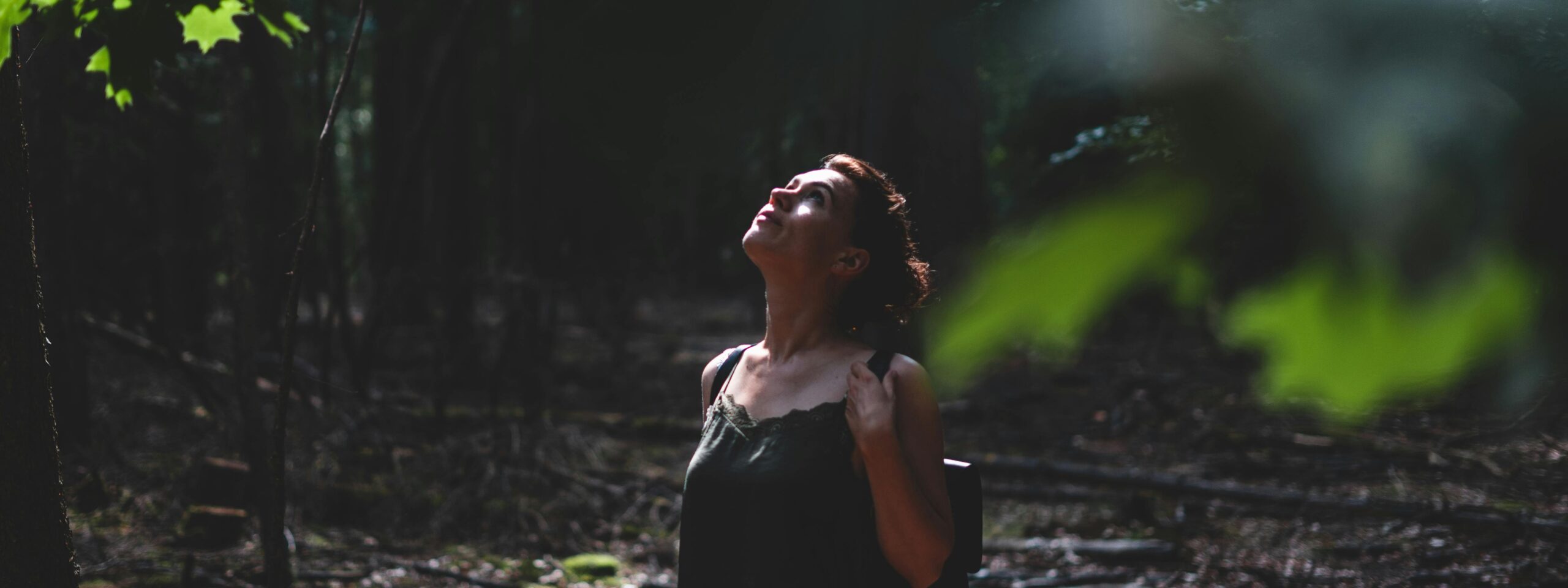 Vrouw kijkt omhoog in een schaduwrijk bos, omgeven door groene bladeren.