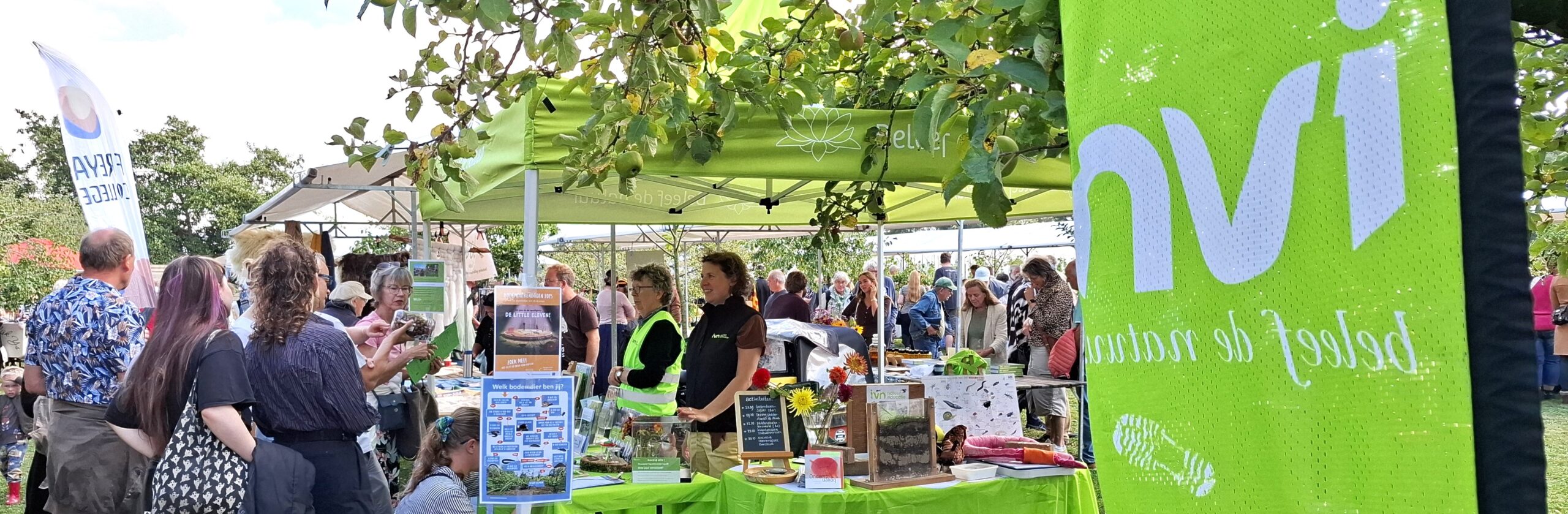 Mensen bij stand met groene tent op een markt, omgeven door bomen en informatiemateriaal.