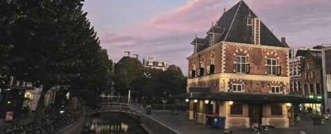 Oud bakstenen gebouw bij een gracht, omgeven door bomen en gebouwen in de schemering.