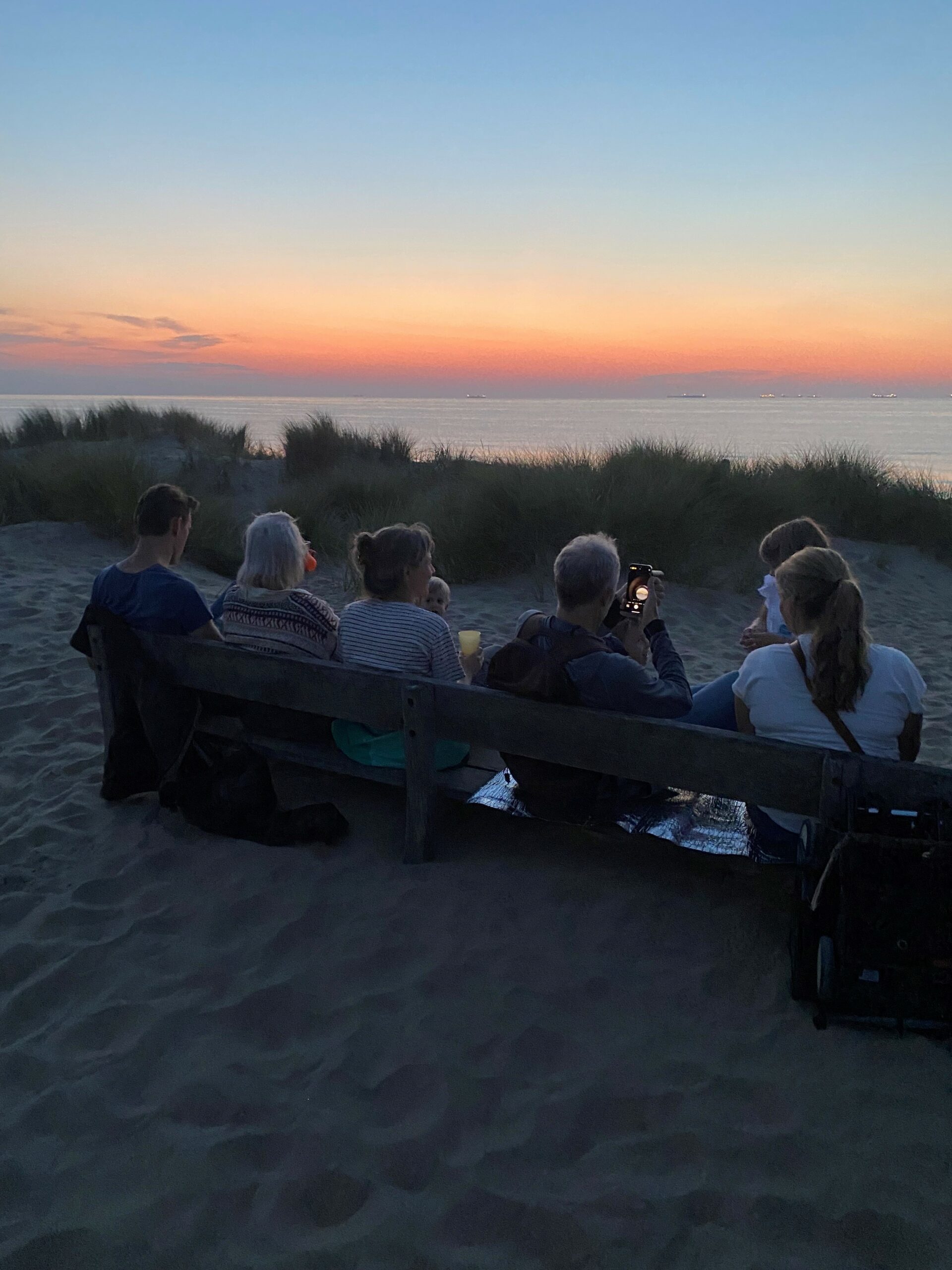 Mensen zitten op een bank in de duinen bij zonsondergang, kijkend naar de zee.