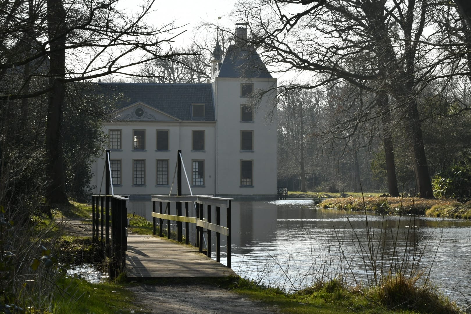 Wit kasteel aan een vijver, brug in de voorgrond, omringd door bomen in een bosrijke omgeving.