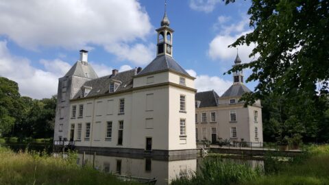 Historisch gebouw met torens en klok, omgeven door bomen en water, onder een blauwe hemel met wolken.