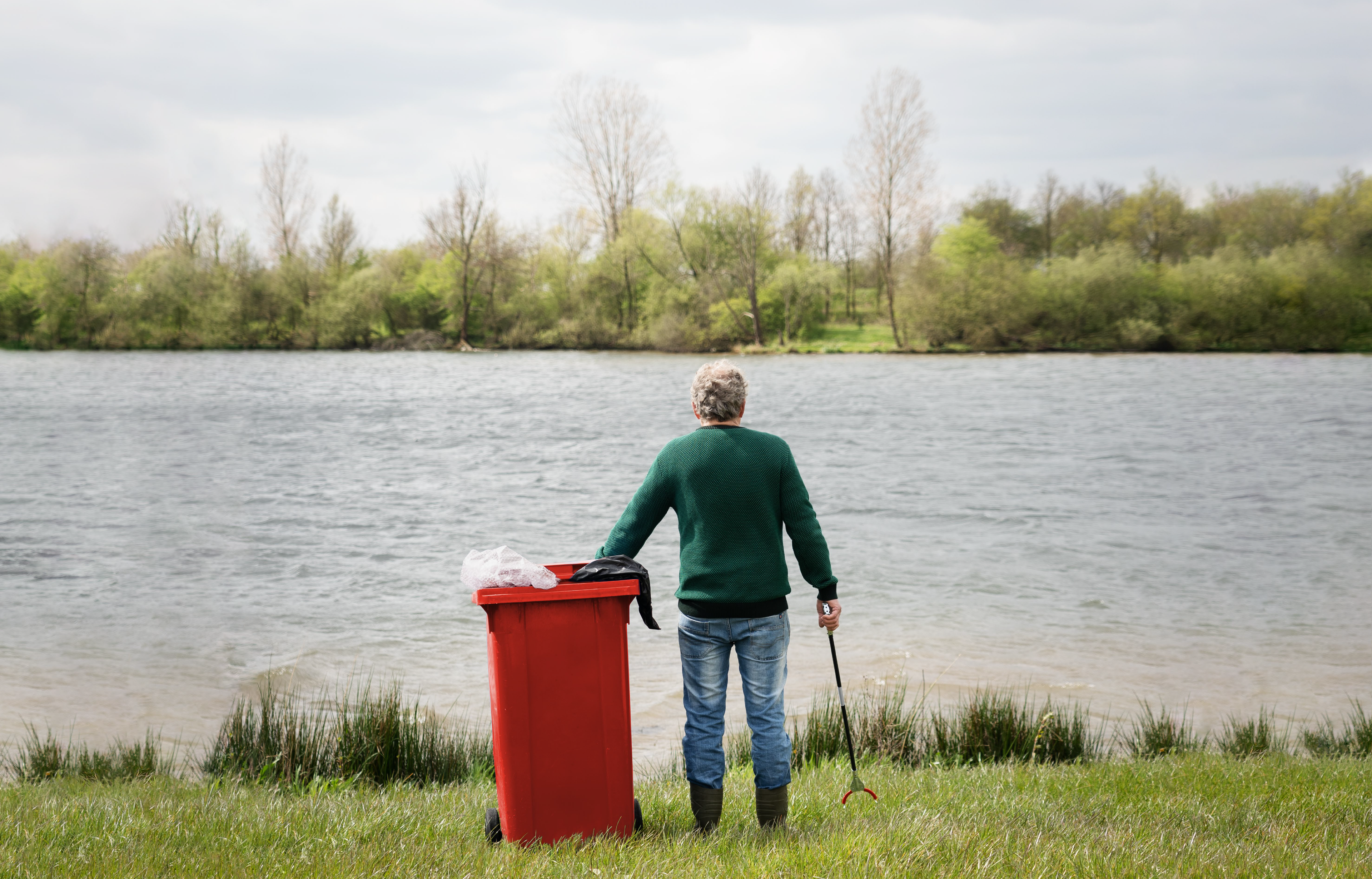 Persoon met grijper bij rode vuilnisbak kijkt uit over water, omgeven door bomen.
