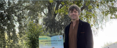 Persoon met prijsbord voor Natuurboekenprijs 2025 in groene, bosrijke omgeving.