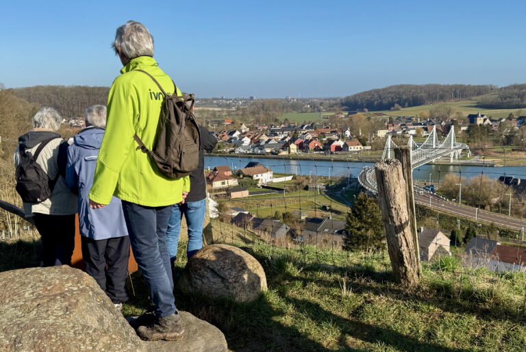 Groep wandelaars kijkt uit over een dorp en brug aan een rivier op een zonnige dag.
