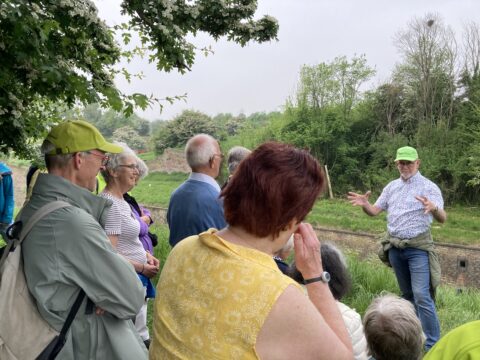 Een gids met een groene pet geeft uitleg aan een groep volwassenen in een groene, bosrijke omgeving.