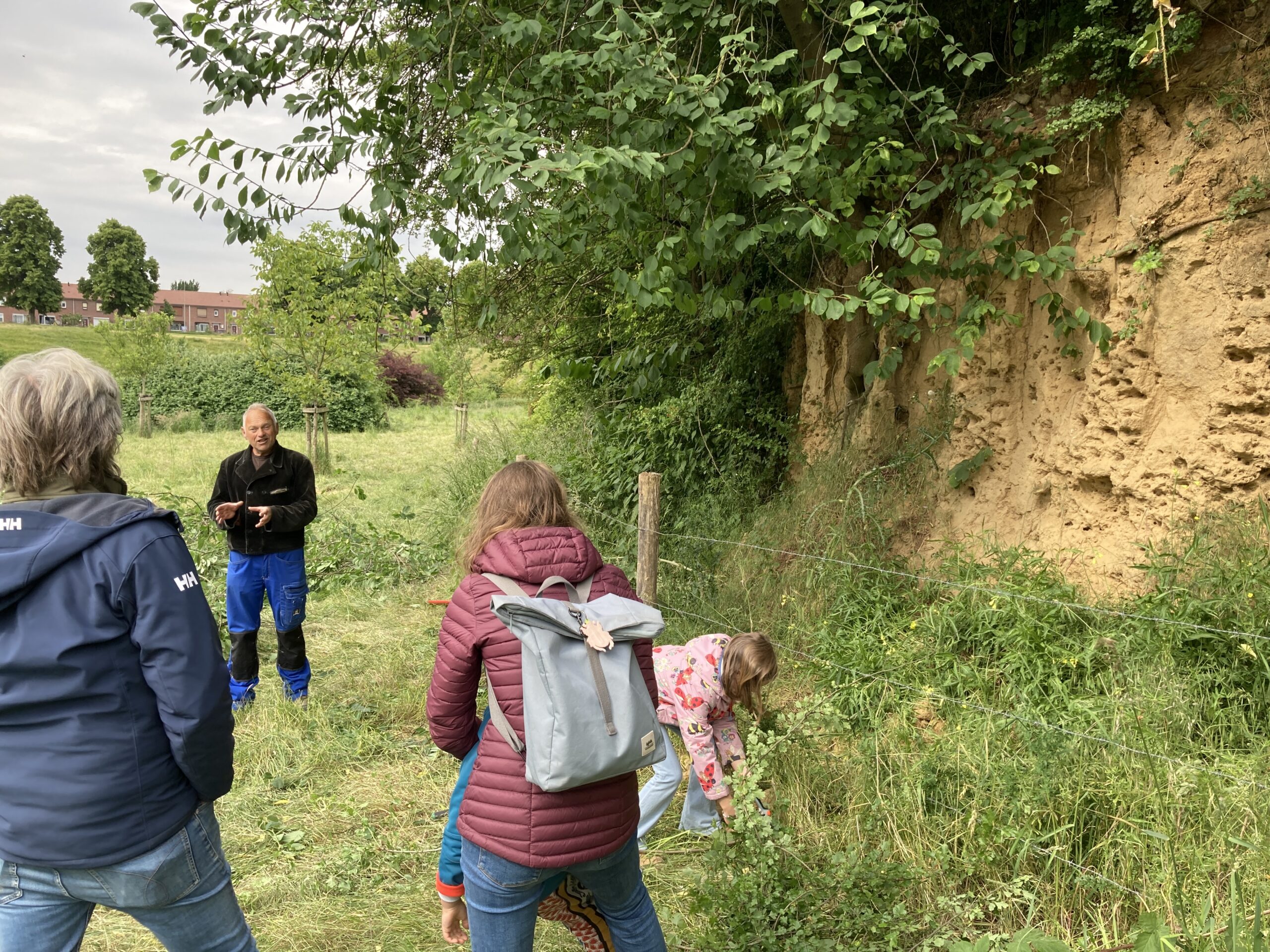 Groep mensen onderzoekt een zandige klif in een groen, landelijk gebied.