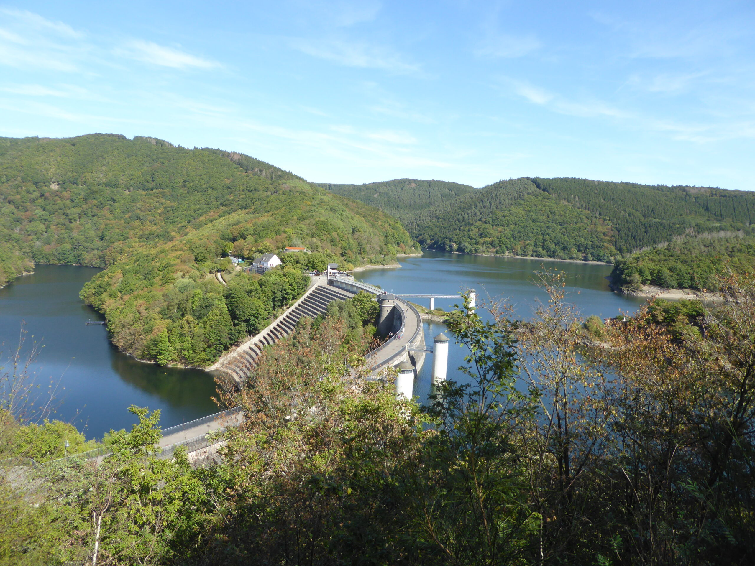 Panoramisch uitzicht op een dam omgeven door groene heuvels en een meer onder een blauwe hemel.