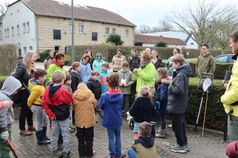 Groep kinderen en volwassenen verzameld buiten, luisterend naar een persoon in een groene jas.