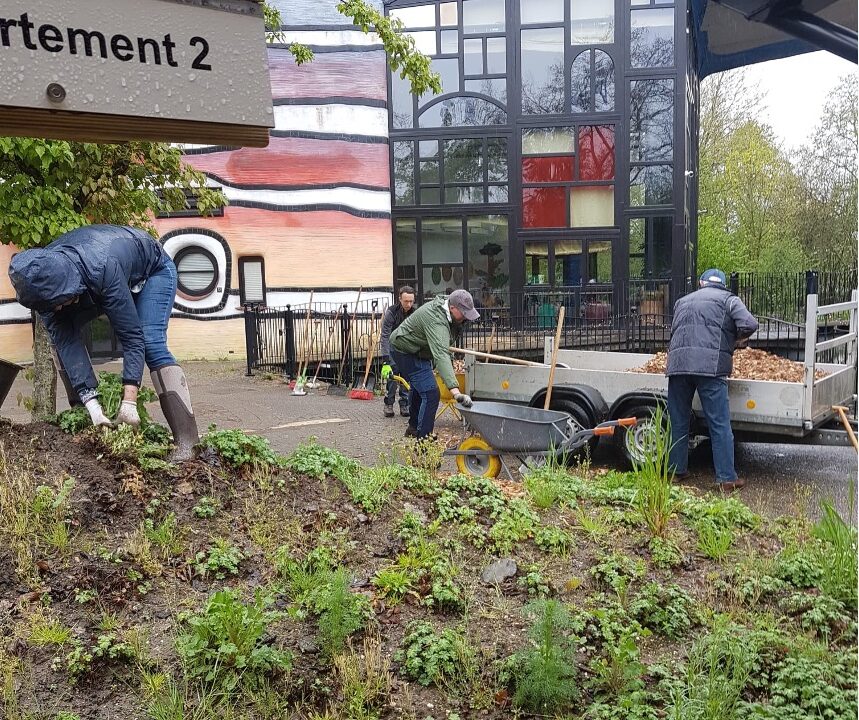 Mensen werken in de tuin met kruiwagens voor een kleurrijk gebouw.