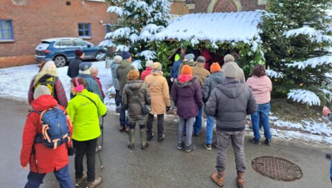 Groep mensen kijkt naar kerststal met sneeuw op daken en bomen, auto op achtergrond.