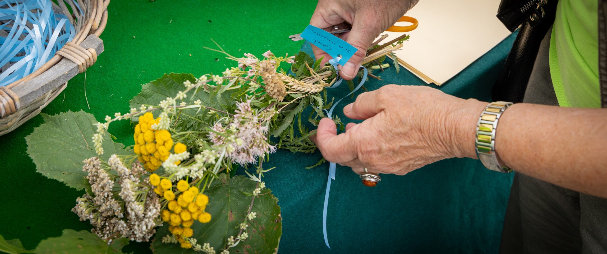 Handen binden bloemen en bladeren met blauw lint op groene tafel.