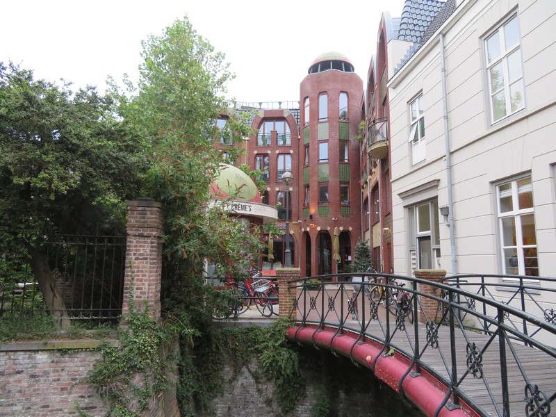 Bakstenen gebouw met torens en balkon, houten brug over gracht, groene bomen aan linkerkant.