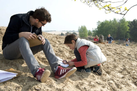 Man en kind onderzoeken zand met vergrootglas, enkele kinderen op de achtergrond.