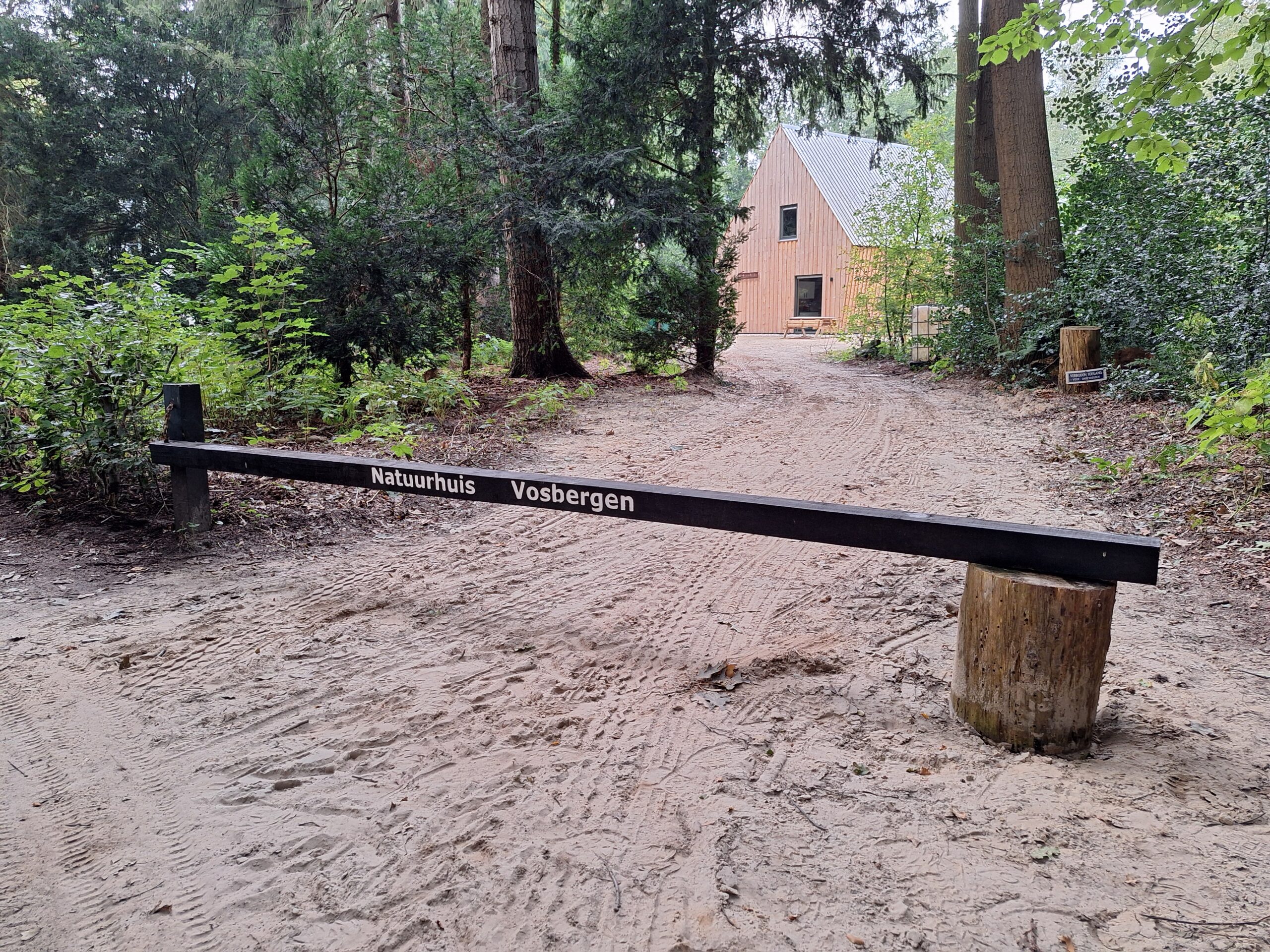 Houten hek bij bospad naar gebouw "Natuurhuis Vosbergen", omgeven door groene bomen.