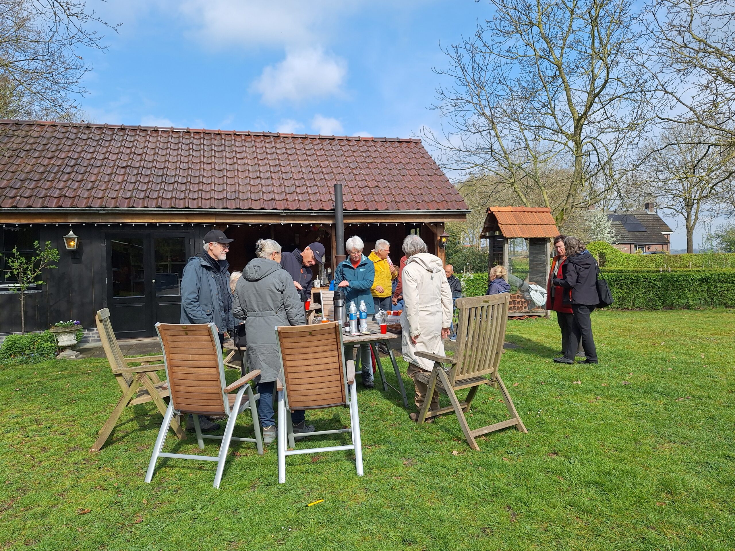 Groep mensen buiten bij houten gebouw en tafel; zonnige dag met gras en bomen.
