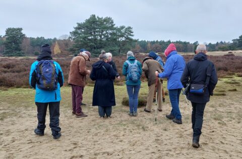 Groep mensen in winterkleding wandelt op een heide, omgeven door bomen.